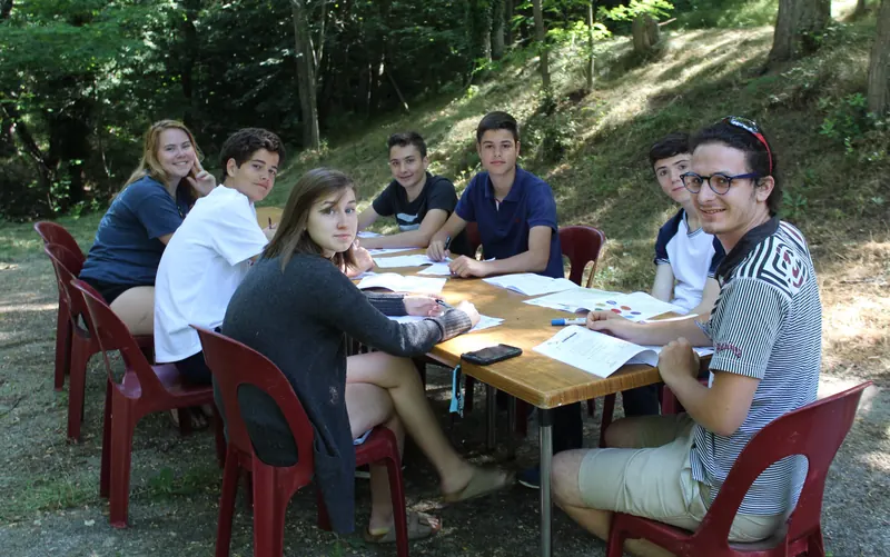 Des jeunes assis autour d’une table en plein air, feuilles et stylos devant eux, souriant à l’objectif