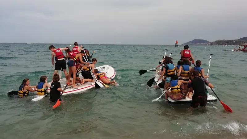 Des jeunes en gilets de sauvetage sur deux très grands paddles en mer, pagaies en main, certains dans l’eau jusqu’à la taille