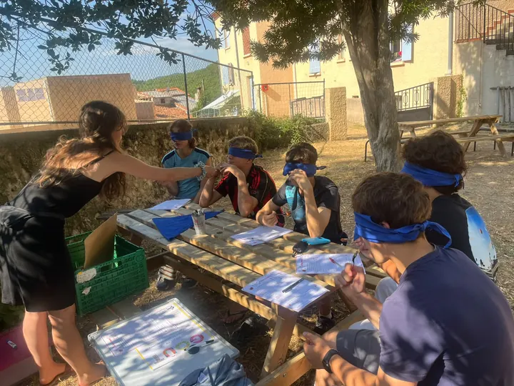 Des jeunes aux yeux bandés autour d’une table de pique-nique, une personne debout leur présente quelque chose à goûter ou à sentir