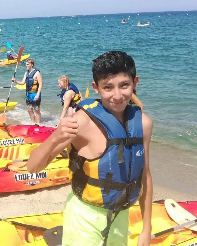 Un jeune souriant en gilet de sauvetage, pouce levé, sur la plage près des kayaks