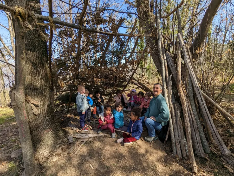 Un grupo de niños y un adulto sentados delante de una cabaña de ramas en el bosque