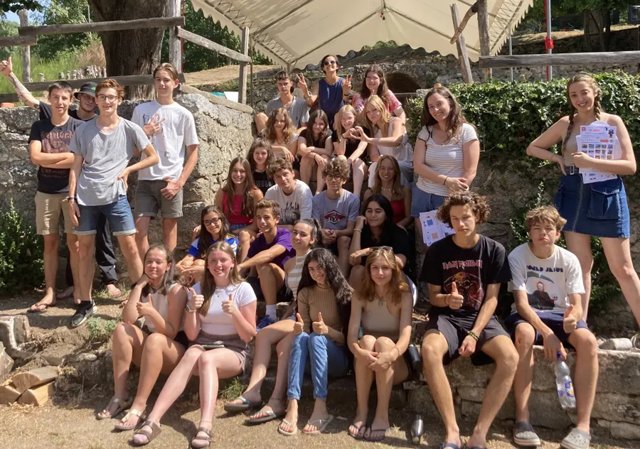 Un gran grupo de alumnos sonrientes sentados en gradas de piedra, a la sombra de un toldo