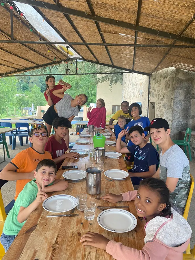 A group of young people sitting at a table under the covered terrace, plates and glasses in front of them, smiling at the camera
