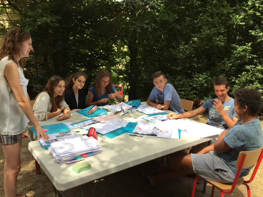 Young people sitting around a table in the woods, with a team leader standing, for a spoken exchange