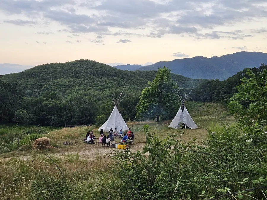 Un groupe de jeunes assis en cercle entre deux tipis, dans une prairie entourée de collines boisées