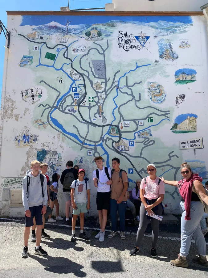 Un groupe de jeunes et deux animatrices devant la grande carte peinte de Saint-Laurent-de-Cerdans, sur le mur d’une maison du village