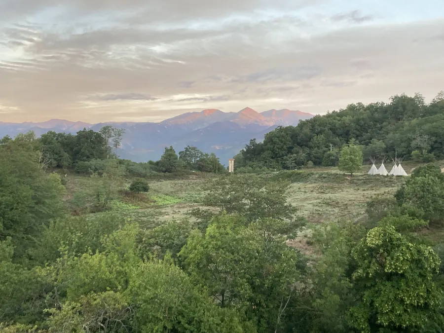 Vue depuis Le Noell : prairie, forêts et tipis, avec le massif du Canigou éclairé de rose au lever du soleil