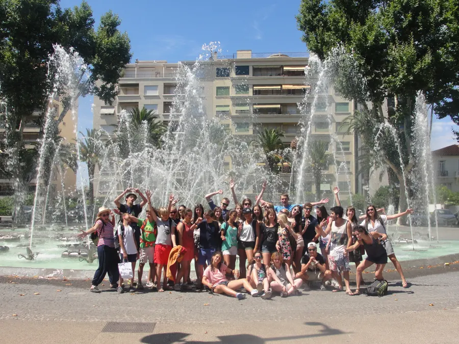 Un grand groupe de jeunes qui lèvent les bras devant une fontaine, sur une place ensoleillée