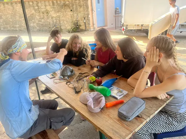 Des jeunes et un adulte autour d’une table en bois couverte d’objets variés : casque, bois de cerf, quilles de jonglage