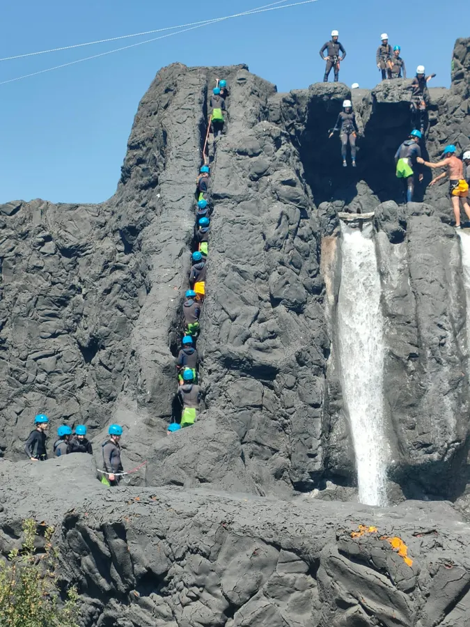 Eine Reihe Jugendlicher mit Helm klettert neben einem künstlichen Wasserfall im Canyoning-Park