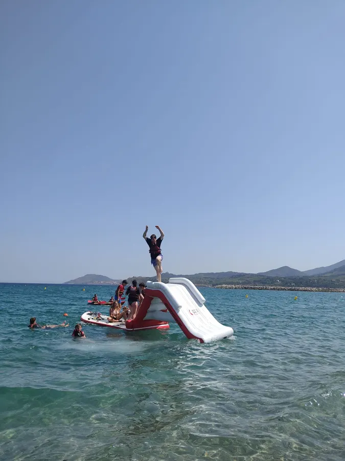 Ein Jugendlicher steht oben auf einer aufblasbaren Rutsche im Meer, andere schwimmen um ihn herum