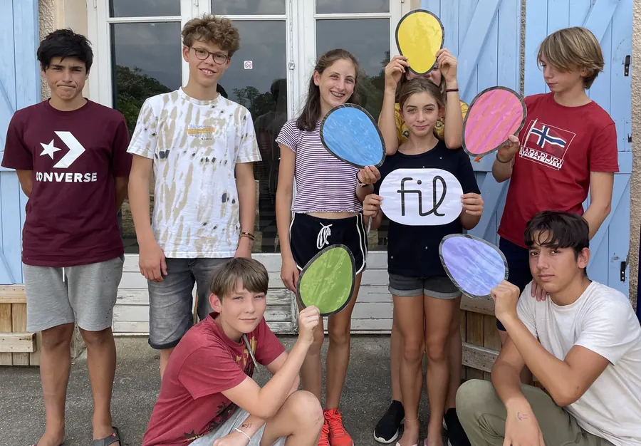 Des jeunes posant devant une maison aux volets bleus, avec de grands pétales colorés et une bulle « FIL » dessinés à la main