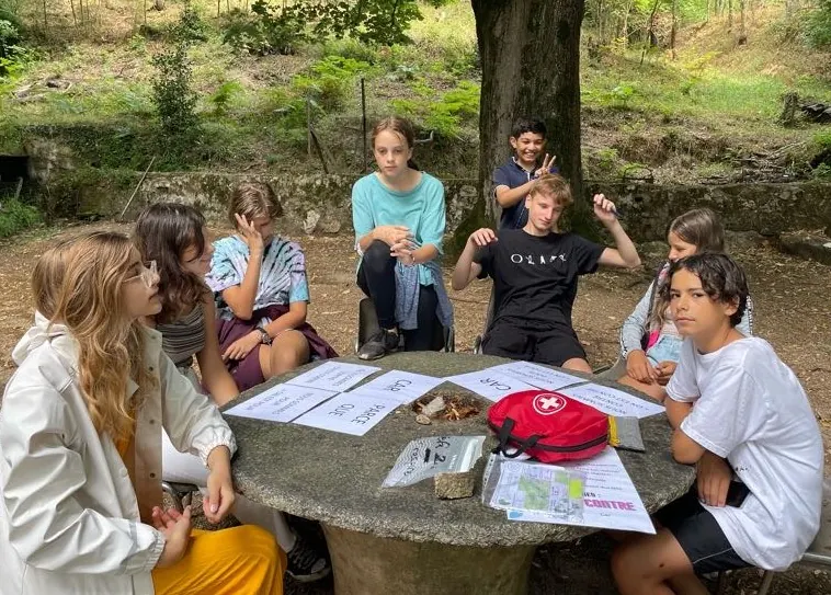 Des jeunes réunis autour d’une table de pierre, sous les arbres, avec des feuilles de jeu et une trousse de secours
