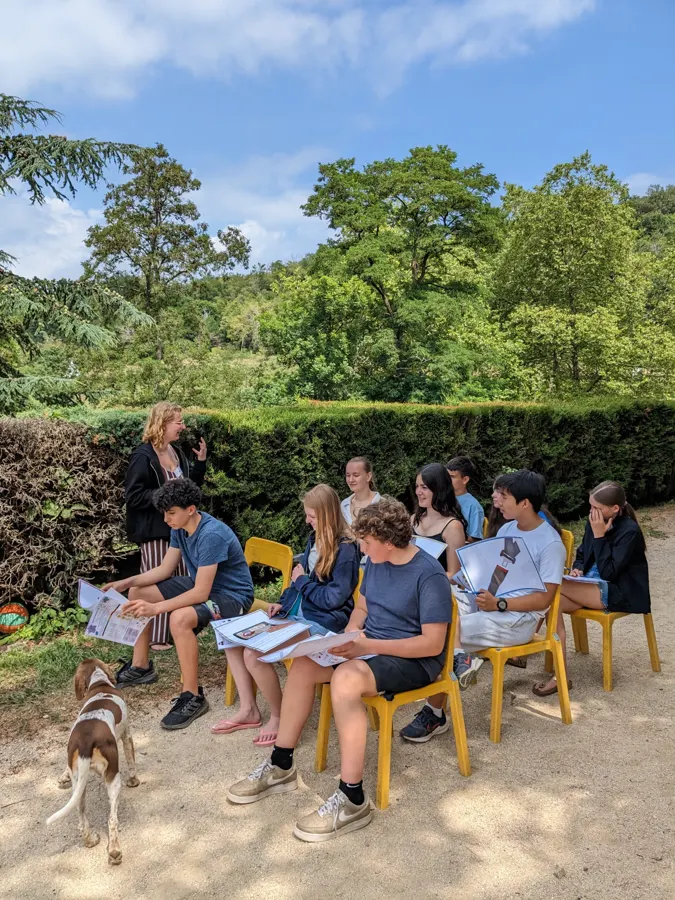 Des jeunes assis sur des chaises en rang dans le jardin, un chien à leurs pieds, devant les arbres