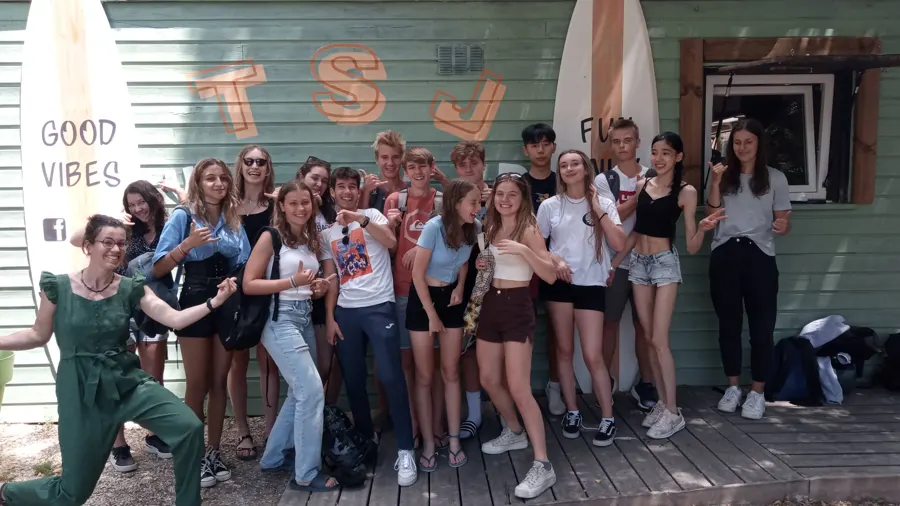Un grand groupe de jeunes joyeux devant le chalet en bois du TSJ Wakepark, avec des planches de wakeboard