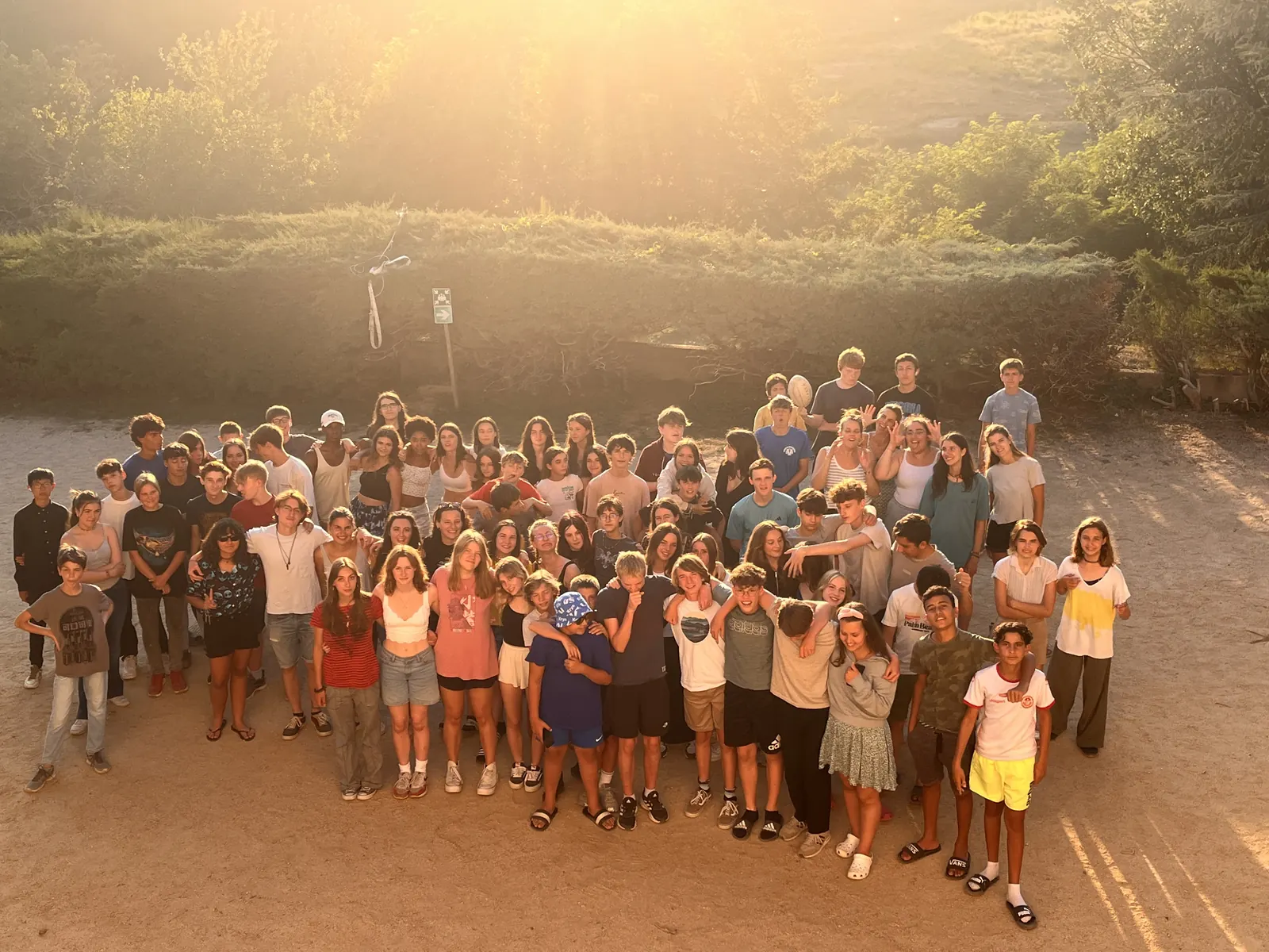 Un grand groupe de jeunes serrés les uns contre les autres, posant ensemble sur une place en terre battue, dans la lumière dorée du soleil couchant