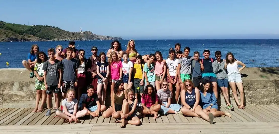 Un grand groupe de jeunes, assis et debout sur une jetée au bord de la mer, avec un phare au loin