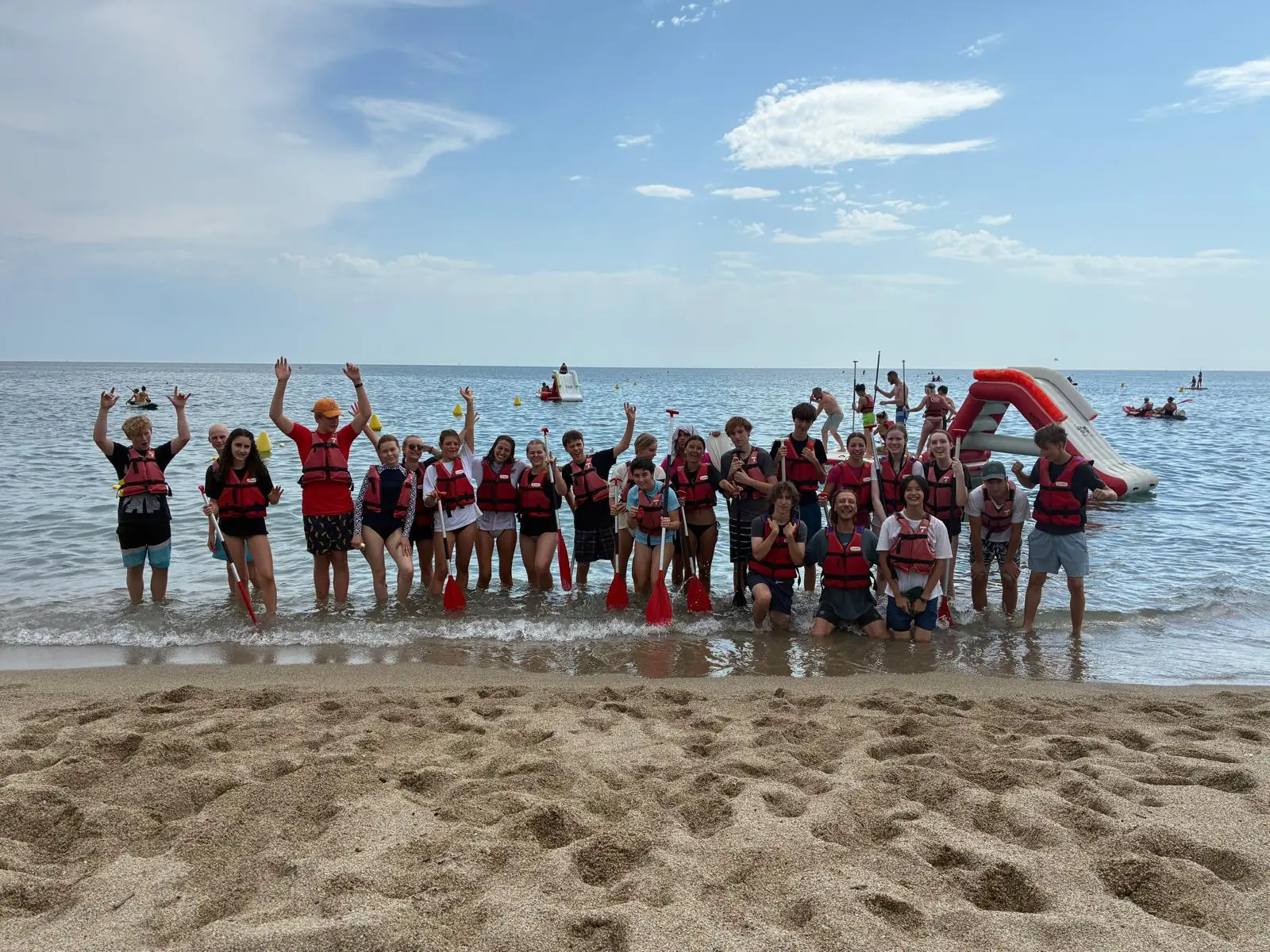 Un groupe de participants en gilets de sauvetage rouges, pagaies à la main, les pieds dans l’eau sur une plage