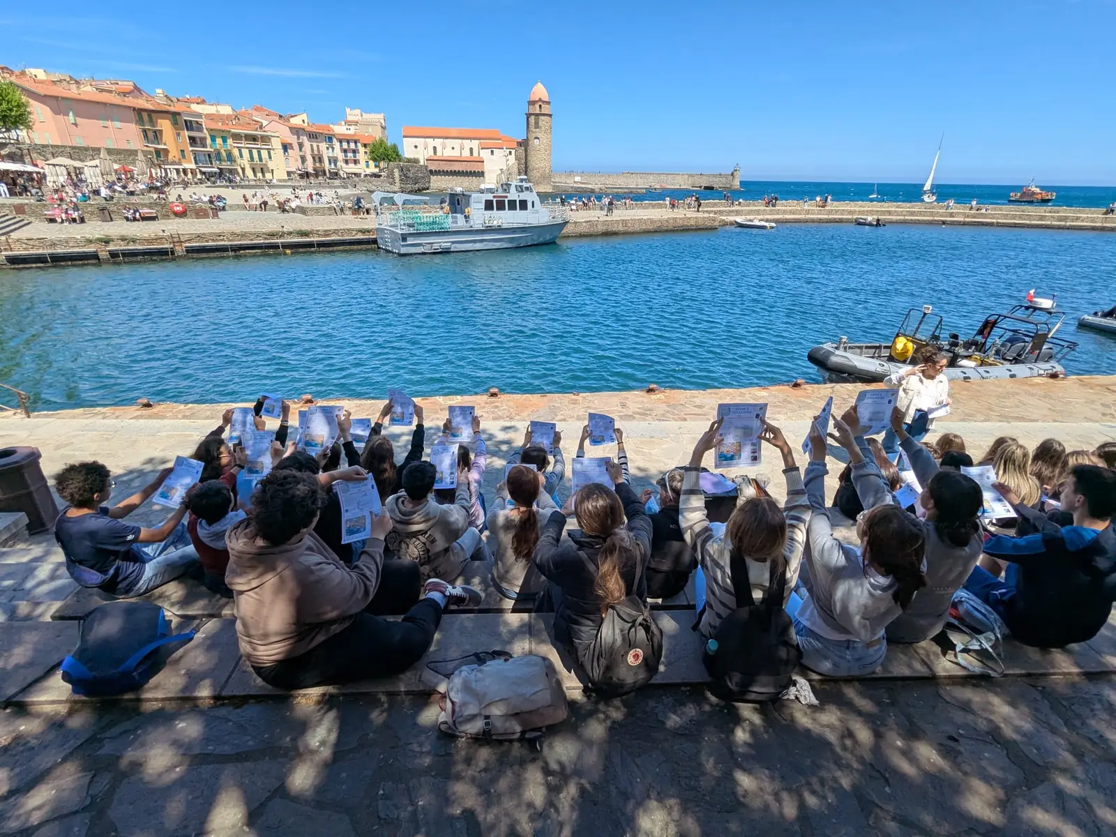 Des participants assis sur le quai du port de Collioure, une feuille de jeu levée devant le clocher et la baie